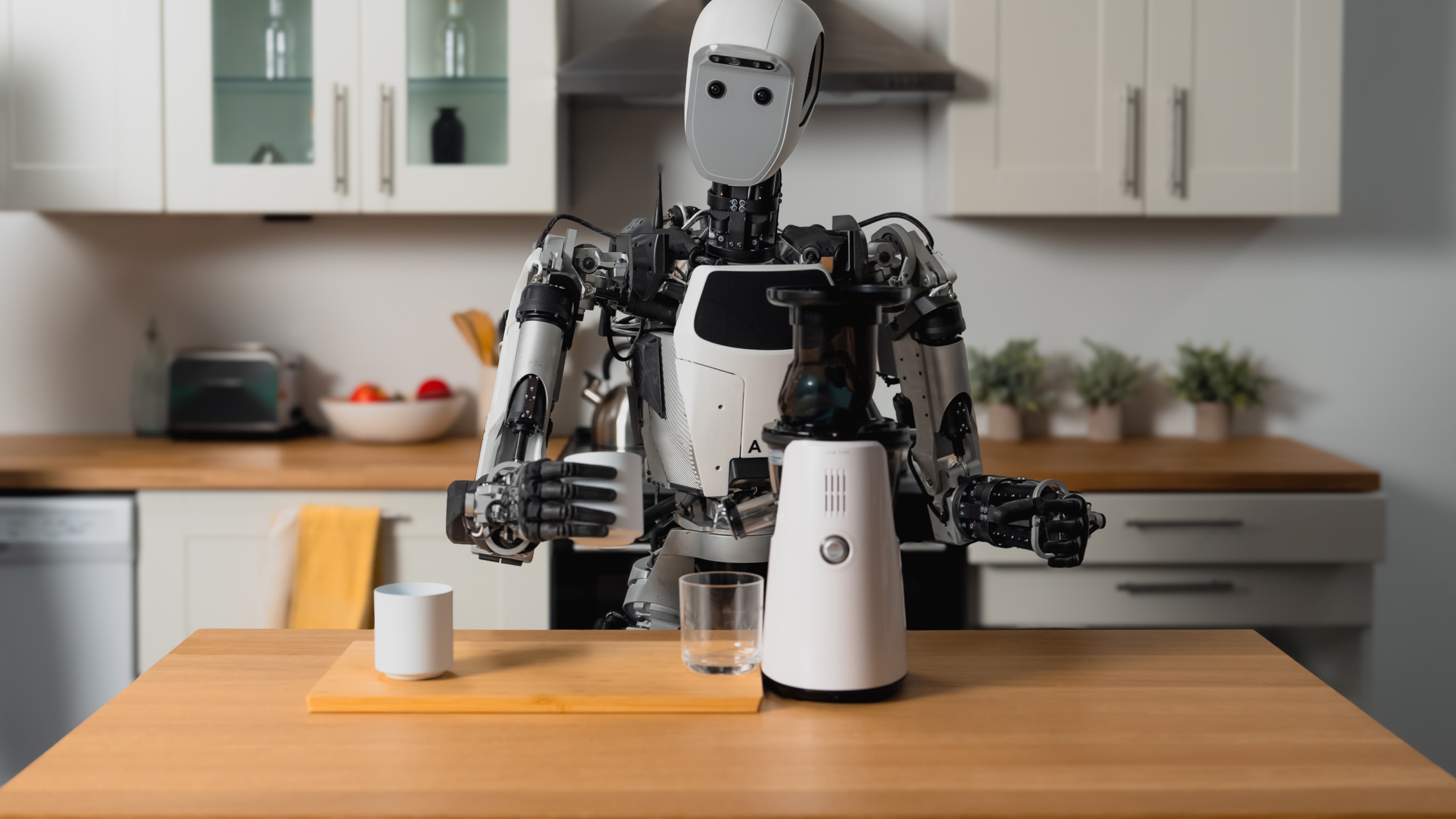 Humanoid robot holding a white mug behind a kitchen counter with a blender, an empty glass, and another mug.
